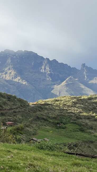 Esta imagen muestra un impresionante paisaje con imponentes montañas, verdes colinas, pequeñas casas y una clara luz solar que ilumina suavemente el escenario natural, creando un apacible ambiente rural.