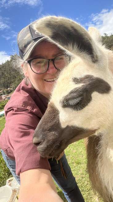 Una persona con gafas y gorra se toma alegremente un selfie con una simpática llama al aire libre, bajo un cielo azul brillante con nubes blancas y vegetación.