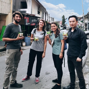 Das Bild zeigt vier Personen, die auf einer Straße stehen und jeweils eine Tüte mit Snacks in der Hand halten. Im Hintergrund sind Gebäude und Fahrzeuge zu sehen, vor einem blauen Himmel mit weißen Wolken.
