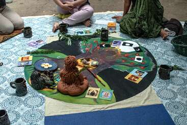Varias personas se sientan con las piernas cruzadas alrededor de un colorido mantel de altar con cartas, tazas, estatuas y objetos naturales, aparentemente participando en una reunión espiritual o ritual sobre una manta.