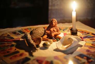 Una vela arde junto a una figura de diosa, rodeada de cartas del tarot, piedras, una concha y salvia, creando un altar místico y espiritual para rituales o meditación.