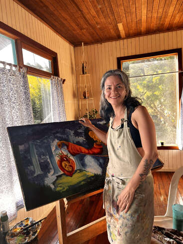 Una mujer sonriente se encuentra en un estudio de arte iluminado por el sol, con un delantal salpicado de pintura, mostrando con orgullo su colorido y surrealista cuadro en un caballete a su lado.