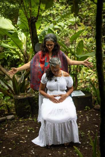 Dos mujeres, vestidas de blanco, se encuentran en un exuberante jardín verde. Una está sentada serenamente, con las manos sobre el regazo, mientras la otra permanece detrás con los brazos abiertos.