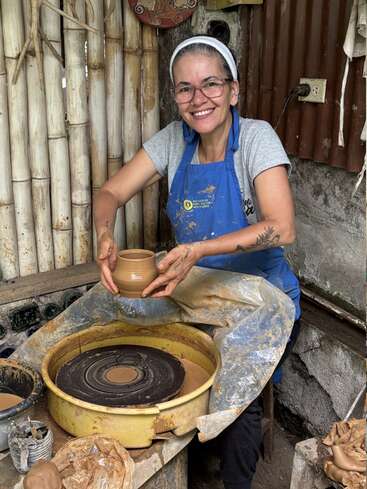 Una mujer sonriente con gafas y delantal azul muestra orgullosa una vasija de barro recién hecha en un torno de alfarería en un rústico y artístico taller.