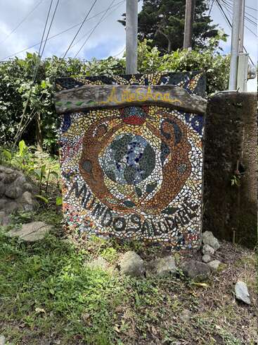 En medio de la vegetación, un colorido cartel de mosaico muestra las palabras "ArteSana" y "Mundos Unidos", con manos artísticas que rodean un globo terráqueo, expresando unidad y creatividad al aire libre.