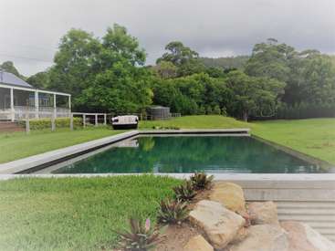 The image depicts a serene backyard scene featuring a rectangular pool, lush green grass, and a house with a covered porch, set against a backdrop of trees and a cloudy sky.