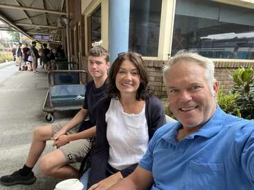 A family of three sits and smiles on a train station bench. The station is busy, with travelers and luggage carts visible in the background. Happy moment.