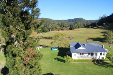 The image depicts a serene rural scene featuring a white house with a blue roof, surrounded by lush greenery and a picturesque landscape under a clear blue sky.