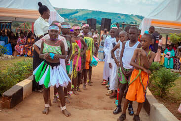A group of children dressed in colorful traditional attire stand outdoors, possibly preparing for a cultural performance. Tents, seated adults, and scenic hills appear in the background.