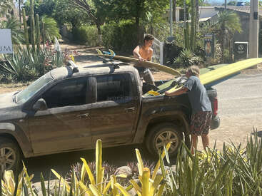 Two young men load long surfboards onto a dusty pickup truck on a sunny day, surrounded by cacti and tropical plants, preparing for an outdoor adventure.