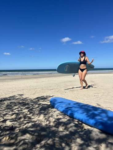 A woman in a black bikini and red hat carries a surfboard on a sandy beach. Another blue surfboard lies in the foreground under tree shade.