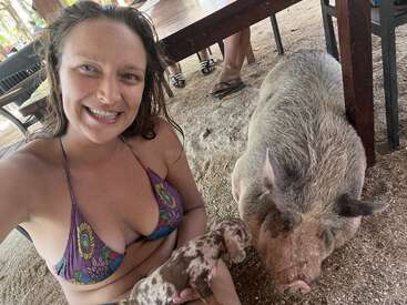 A smiling woman in a colorful bikini takes a selfie with a large pig and a small piglet under a wooden table on a sandy surface outdoors.