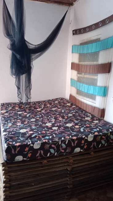 Se trata de una habitación sencilla que cuenta con una cama con un colchón estampado de flores sobre un somier de madera, un mosquitero negro colgado y una ventana con cortinas de rayas de colores.