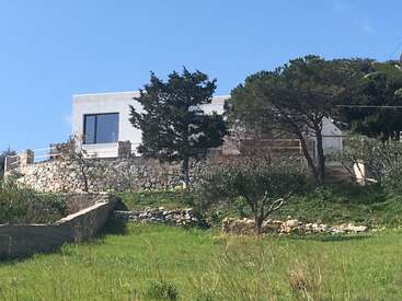 The image depicts a modern white house with a stone wall and a grassy yard, featuring trees and a clear blue sky in the background.