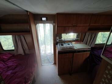 Esta imagem mostra o interior de uma van de camping com armários de madeira, uma cozinha compacta, fogão a gás, camas dos dois lados, janelas com cortinas e luz natural.