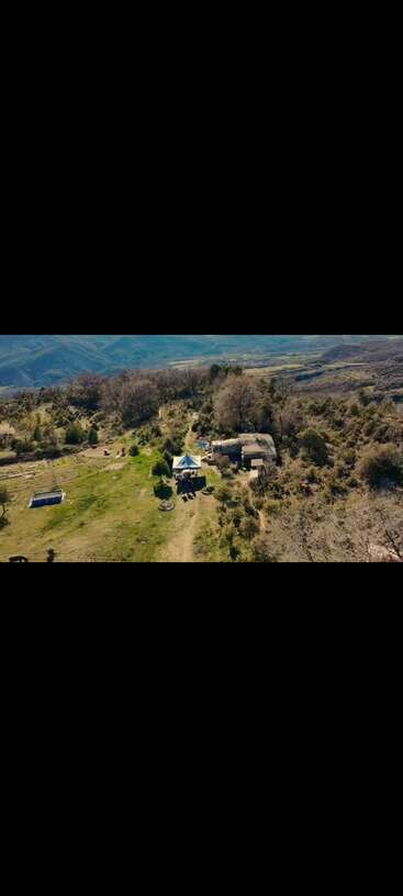 Essa imagem mostra uma área rural isolada com uma casinha, uma barraca, árvores espalhadas, um terreno gramado, uma piscina, montanhas e um céu azul claro ao fundo.