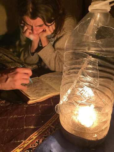 Eine Frau studiert konzentriert neben einem Notebook, das von einer selbstgebastelten Laterne aus einer Plastikflasche und einer Kerze beleuchtet wird, was eine warme, gemütliche Atmosphäre schafft.