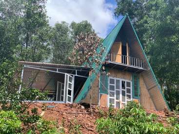 Esta imagen muestra una casa de estructura en A con tejado verde, enclavada entre árboles. La casa tiene grandes ventanales, un balcón superior y una ladera inclinada.