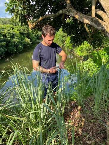 Un hombre está de pie junto a la orilla de un frondoso río rodeado de hierba alta y árboles. Parece concentrado, posiblemente examinando o preparando algo entre sus manos, disfrutando de la naturaleza.