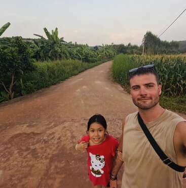 Un hombre y una niña de pie en un camino rural rodeado de exuberante vegetación, sonríen a la cámara. La niña lleva una camiseta roja de Hello Kitty.