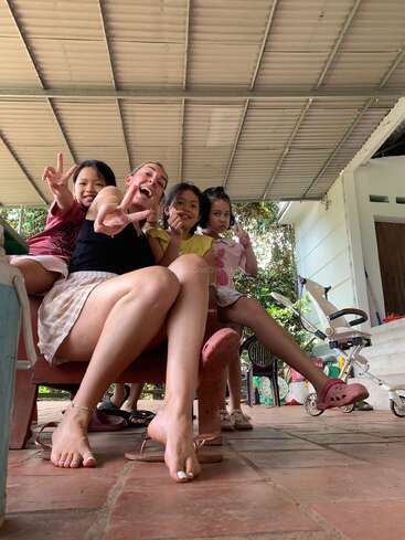 Cuatro personas, un adulto y tres niños, se sientan juntas en un banco haciendo signos de la paz, sonriendo y disfrutando de un momento relajado y alegre en un patio cubierto.