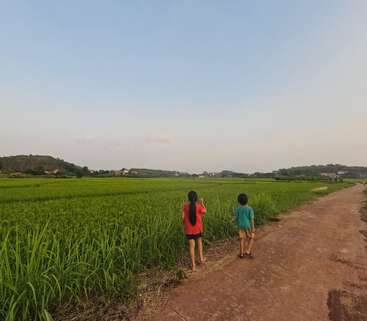 Dos niños caminan por un sendero de tierra junto a frondosos campos verdes bajo un cielo inmenso y despejado. Casas lejanas y suaves colinas completan la apacible escena rural.