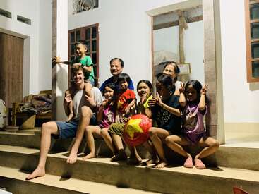 Un grupo de personas, entre las que hay adultos y niños, se sientan alegremente en las escaleras de una casa por la noche, sonríen, posan y sostienen una pelota roja con forma de estrella.