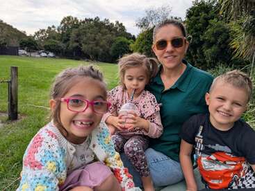 Una mujer y tres niños sonrientes disfrutan de un picnic sobre hierba verde, con árboles a sus espaldas. Un niño sorbe una bebida, todos parecen felices al aire libre.