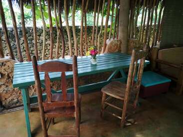 A rustic outdoor seating area features a turquoise wooden table, two weathered chairs, a small flower vase centerpiece, and a backdrop of natural wooden fencing.