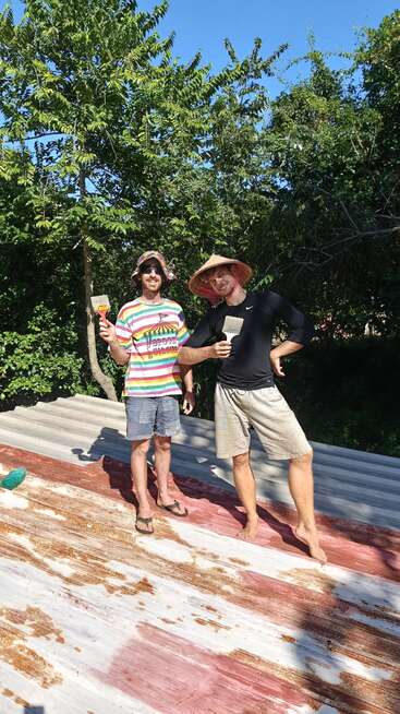 Two men standing barefoot on a roof, holding paintbrushes, smiling under the sun. Both wear hats and casual clothes, surrounded by green trees and blue sky.