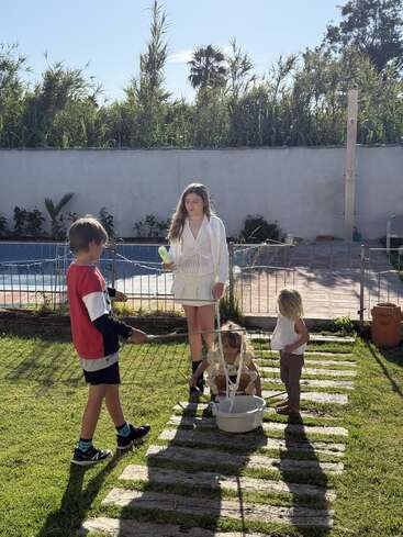 Cuatro niños juegan juntos al aire libre en un patio soleado. Están haciendo pompas gigantes con un cubo y una cuerda. Están rodeados de una exuberante vegetación y una valla.