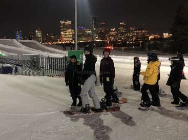 L'image représente un groupe de personnes sur des snowboards dans un environnement urbain de nuit, avec la ligne d'horizon de la ville à l'arrière-plan et une colline enneigée à gauche.