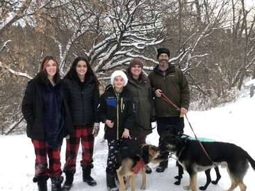 L'image représente une famille de cinq personnes, dont deux adultes et trois enfants, posant avec leurs deux chiens dans un décor hivernal enneigé, entouré d'arbres et de buissons.