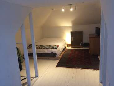 Dieses Bild zeigt ein gemütliches Schlafzimmer im Dachgeschoss mit weißen Wänden und Böden, einem niedrigen Bett, Holzmöbeln, einem roten Teppich und warmer, sanfter Beleuchtung.