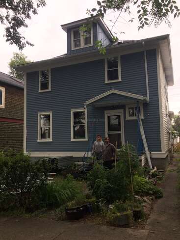 Ein zweistöckiges blaues Haus mit weißen Zierleisten und einer kleinen Dachgaube im Obergeschoss. Zwei Personen stehen auf der Eingangstreppe, umgeben von üppigen grünen Gartenpflanzen.