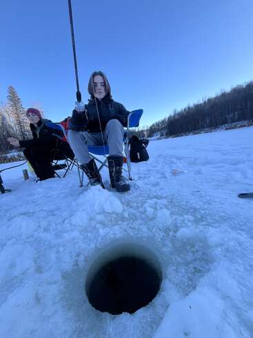 Zwei Menschen sitzen auf einem zugefrorenen See und angeln durch ein rundes Loch im Eis. Warm angezogen konzentrieren sie sich auf ihre Angelruten. Um sie herum liegen Schnee und Bäume.