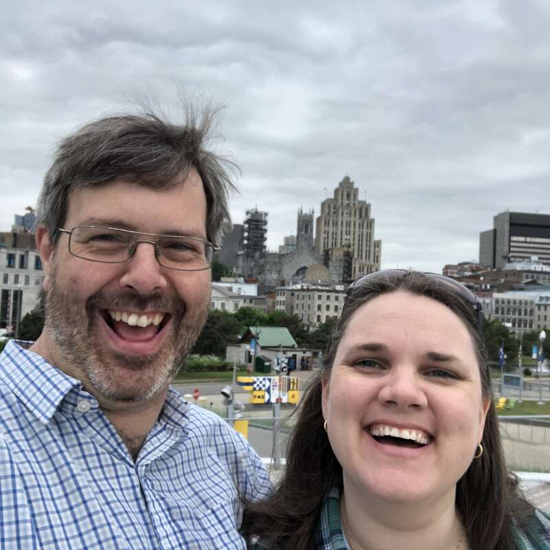 Ein lächelndes Paar macht draußen ein fröhliches Selfie. Hinter ihnen bildet eine Stadtlandschaft mit historischen Gebäuden und bewölktem Himmel eine lebhafte Kulisse.