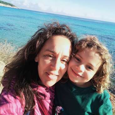 La imagen muestra a una mujer y un niño posando para un selfie frente a una masa de agua, con el sol brillando intensamente sobre sus rostros.