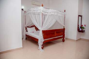 La imagen muestra un dormitorio limpio y minimalista con una cama de madera con dosel envuelta en una mosquitera blanca, paredes blancas, suelo de baldosas y un pequeño estante con espejo.