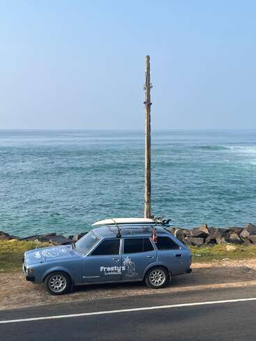 Ein altes blaues Auto mit Surfbrettern auf dem Dach steht am Meer. Auf dem Auto steht „Frostys“. Im Hintergrund sieht man einen Strommast, Wellen und Surfer.