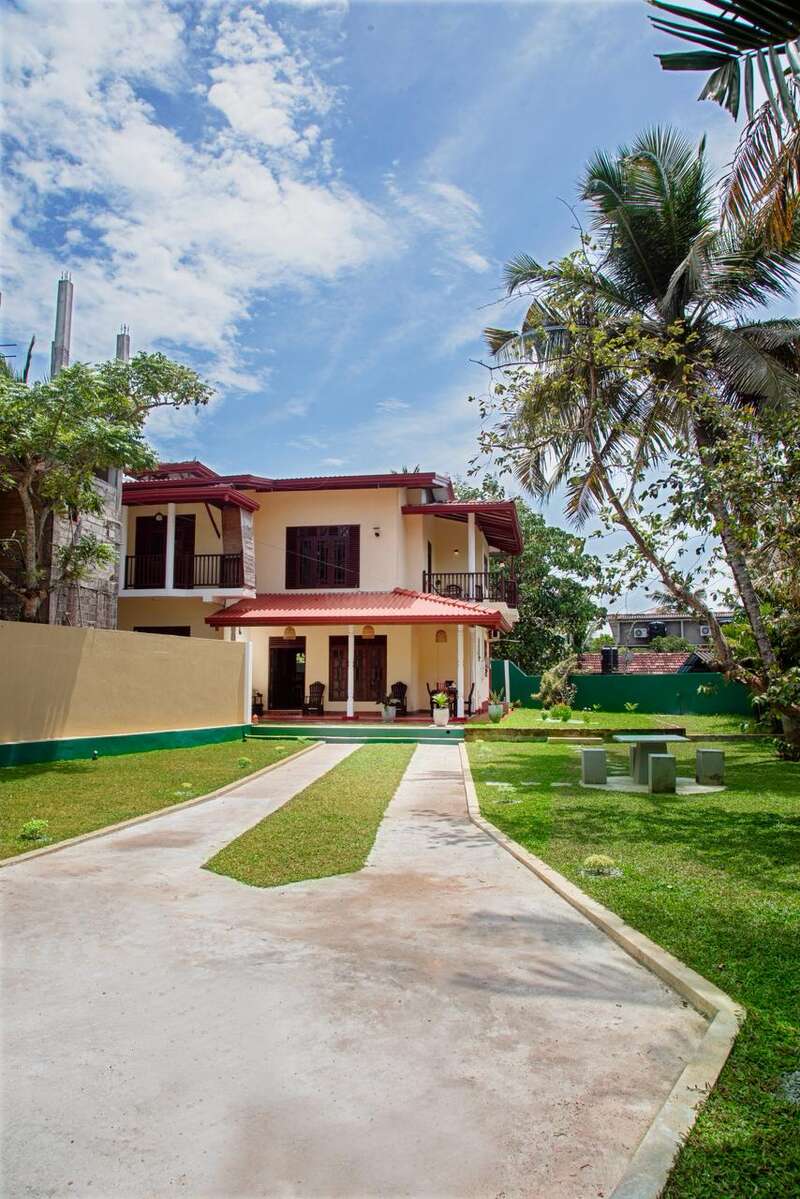 Una casa de dos plantas con tejado rojo y paredes amarillas, rodeada de un patio verde, palmeras, camino de entrada, cielo azul y zona para sentarse al aire libre.