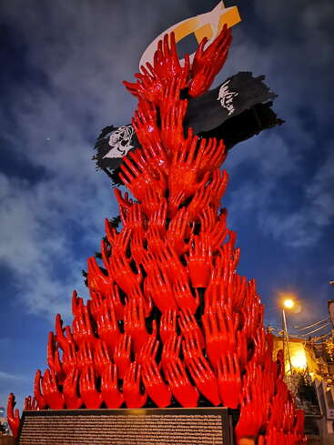 Uma escultura impressionante feita de vários ponteiros vermelhos que se estendem para cima, em um cenário dramático de céu noturno. No topo, você pode ver o formato de uma estrela e uma lua crescente.