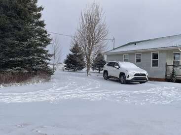 Un SUV blanc recouvert de neige est garé à côté d'une maison. La neige recouvre le sol, les arbres et les environs, créant une scène rurale paisible et hivernale.