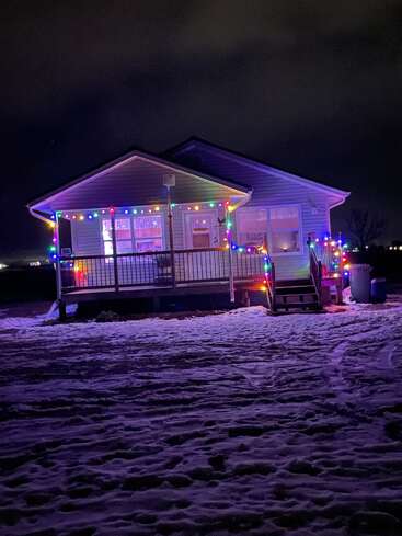 Une petite maison décorée de lumières de Noël colorées brille chaleureusement la nuit. La neige recouvre le sol, créant une atmosphère festive et chaleureuse de vacances d'hiver.