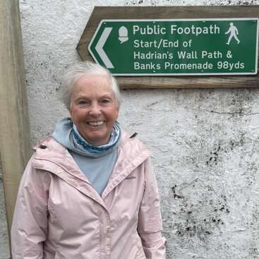 Eine ältere Frau in einer hellrosa Jacke lächelt vor einem Schild, das den Anfang oder das Ende des Hadrian’s Wall Path und der Banks Promenade markiert.