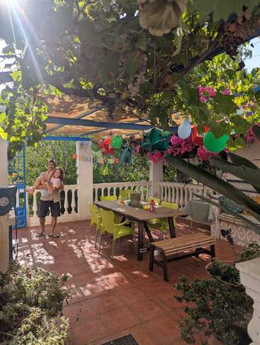Un patio soleado decorado para una celebración, con globos de colores, una pancarta de "Feliz cumpleaños", follaje verde y dos personas felizmente abrazadas bajo el toldo de parra.