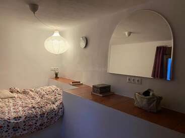 Un dormitorio acogedor y minimalista presenta una cama con sábanas de colores, un gran espejo arqueado, un farol colgante, libros, una planta y una cortina morada.