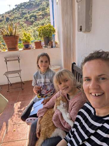 Una mujer y dos niños sentados en un porche con plantas en macetas, una hermosa vista de la ladera y un relajado gato naranja y blanco en su regazo, sonriendo.