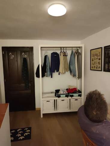 Una habitación acogedora con un armario, ropa colgada, cestas debajo y algunas maletas. Una persona sentada en primer plano, arte decora las paredes. Iluminación cálida.