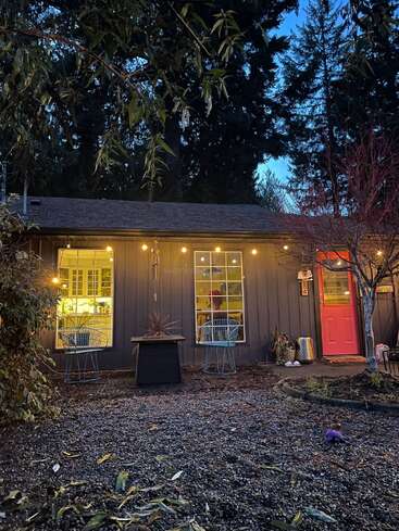 A cozy cottage with warm glowing lights, two blue chairs, a red door, and large windows is nestled among trees as evening falls, creating inviting charm.
